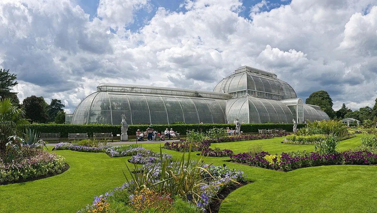 De victoriaanse Palm House in Kew Gardens weerspiegeld in het vijverwater, omgeven door groen en blauwe lucht